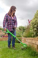 Woman using strimmer to cut long grass along edge of raised bed