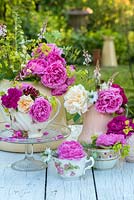 Cut roses arranged in enamel tea set, against a garden backdrop.
