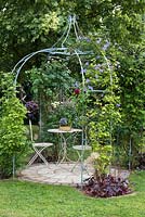 Ornate metal gazebo or arbour above a circular patio with table and chairs. Climbers such as 
 Clematis 'Blue Angel' are trained up outside.

