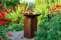 Bird bath of oak sleeper pedestal and mild steel bowl surrounded by Crocosmia 'Lucifer at  Veddw House Garden, Monmouthshire, Wales, UK.
