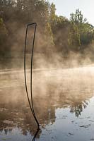Mist rising from the lake with metal sculpture at sunrise. Plaz Metaxu Garden, Devon, UK.
