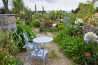 Sheltered gravelled terrace with seating surrounded by cordylines, olives and agapanthus with scarlet pelargoniums in pots on ledge beyond