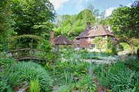 View of wooden bridge and 16th century cottage through spring planting. Copyhold Hollow, Sussex, UK. 