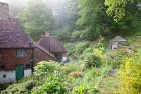 View of cottage and greenhouse surrounded by spring woodland garden. Copyhold Hollow, Sussex, UK. 