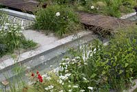 A rill edged with white Laceflower, Orlaya grandiflora, salvias, fennel and grasses, with a wooden bridge crossing the water. The Style and Design Garden, designed by Ula Maria, sponsored by London Mosaic CED Garden Brocante Online, RHS Hampton Court Palace Garden Show, 2018.