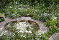 Circular pool edged with a cobbled path and herbs. The Health and Wellbeing Garden, designed by Alexandra Noble, sponsored by CED Ltd, Majestic Trees, Marshal Murray, Hampton Court Palace Flower Show, 2018 