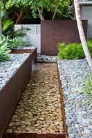 Corten steel water feature with pebbles or cobbles, block planting of Osmoxylon lineare - Migros Bush - near steel panel

