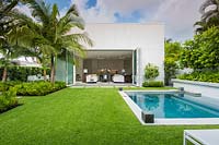 View over Zoysia grass lawn to back of house with folding doors, on one side a swimming pool and on the other beds of foliage plants including palms