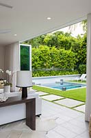 View from the sitting room to the garden showing folding doors opening out on to white slabs set in Zoysia grass, with swimming pool and raised bed of Citrus - Orange trees, under planted with Aechmea - Bromeliad, beyond

