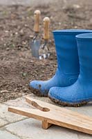 Muddy boots and boot Jack on paving with soil and hand tools beyond