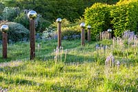 View of meadow with Camassia subsp. leichtlinii and avenue of 
stainless steel mirror globes mounted on wooden posts casting shadows
