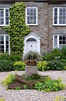 Graveled area with central stone plinth and potted plant, with view to front door surrounded by perennial borders. Bosvigo House, Cornwall, UK.