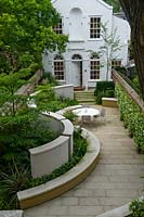 View of private urban back garden and house, with pale paving and curved bench and wall. 