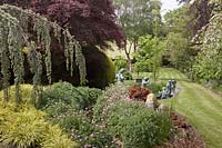 View across herbaceous border to lawn with bronze figures of children in a rill. Planting includes Carex, Cedrus, Acers and Geraniums 