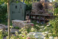 A romantic country garden planted with mostly medicinal plants, a shepherd's hut, a curving stone bench, and an eclectic display of objects. The Naturecraft Garden, designed by Pollyanna Wilkinson, RHS Hampton Court Palace Garden Festival, 2019.