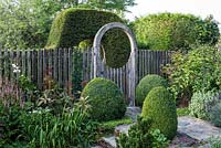 An oak moongate set in a tall picket fence, with a clipped yew arch behind. Box balls flank the stone path.