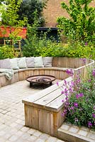 Seating area with fire pit and round wooden bench surrounded by Erysimum 'Bowles's Mauve', Prunus avium 'Stella' and Cornus alba 'Sibirica' 