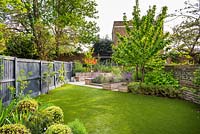 View across the garden with artificial grass to seating area with fire pit and round wooden bench surrounded by Erysimum 'Bowles's Mauve', Prunus avium 'Stella', Rubus idaeus and raised vegetable beds. 
