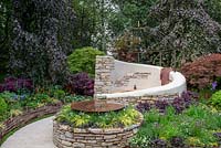 Fagus sylvatica 'Purple Mountain' trees providing the backdrop to a dry stone wall seating area with decorative sundial dome and circular pool, plants include Loropetalum, Acer palmatum, Carex and Erigeron karvinskianus - Miles Stone: The Kingston Maurward Garden, RHS Chelsea Flower Show 2019.