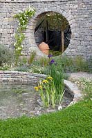The Harmonious Garden of Life, the circular pond is planted with Iris pseudacorus and Iris laevigata, its shape is echoed in the limestone wall with an oculus through which we can see a dome-topped terracotta bee hive, the lawn at the front is planted with clover - Sponsors: Mr and Mrs Cawthorn, Margheriti Piante.