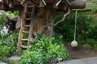The RHS Back to Nature Garden, view of the treehouse with its ladder and rope, it is underplanted with ferns, Dryopteris filix-mas and Athyrium filix-femina,  Luzula nivea, Lamium and Vinca. RHS Chelsea Flower Show 2019.