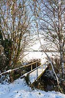 View out of garden over bridge to the fields beyond, covered in snow. Including alder trees 'Alnus glutinosa' in the foreground.