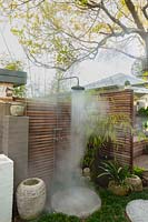 An outdoor shower, showing hot water coming out of it, attached to a hardwood timber privacy screen, with potted bamboo, pots and round stepping stones.
