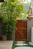 A clump of bamboo growing in the back corner of a garden next to a bespoke hardwood timber gate showing bespoke round stepping stones path, interplanted with Mini mondo grass, Ophiopogon japonicus Nanus, featuring a carved Indonesian pot.
