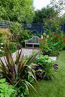 Contemporary  garden in West London with artificial lawn - view through borders with Phornium Queen, Helenium Moerheim Beauty, Hydrangea Vanille Fraise, Acer Katsura, Prunus Autumnalis Rosea towards stone patio with a wooden seat.