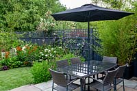 Contemporary garden in West London - view from stone paving patio with table and parasol through borders with Verbena bonariensis, Echinacea Magnus Superior, Helenium Moerheim Beauty, towards artificial lawn.