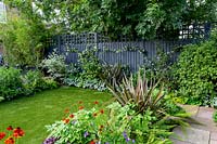 Contemporary garden in West London with artificial lawn and grey painted fence with trellis - view through borders with Phornium Queen, Helenium Moerheim Beauty, Geranium Rozanne, Polystichum setiferum Herranhausen.