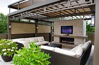 View of outdoor room on a roof garden, showing relaxation area of sofas, flat screen television and outdoor fire pit. All sheltered by metal pergola with wall lights 