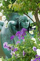 View past Euonymus, underplanted with flowering Petunia and Verbena to containerised statue of woman. 