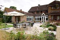 View across patio with small beds of perennials and dining area, towards back of the house with patio doors and brick extension
