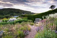 View over prairie-style planting a bench backed by a hedge, wooded hills beyond. Plants include: Molinia caerulea subsp. caerulea 'Poul Petersen', Achillea, Hordeum jubatum 'Early Pink' and Calamintha nepeta subsp. nepeta 'Blue Cloud' with white Erigeron annuus 