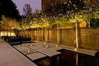 Row of standard trees, paving and water, lit up with spotlights in contemporary, city garden. 