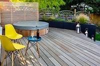 Contemporary garden in Wimbledon with decking on two levels with yellow chairs and table with wine bottle. Wooden steps with lanterns connect the two deck levels. With grey raised bed with perennials. In the background is a hot tub.