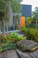 A hardwood timber deck next to a pond with a sandstone boulder insert and a lush, dense planting of colourful Bromeliads, and Dwarf papyrus plants featuring a Queensland Bottel tree festooned with air plants and Spanish Moss.