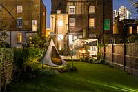 View across illuminated garden with cacoon hanging tent to the house, outdoor kitchen and dining area.  