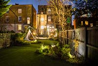 View across illuminated garden with cacoon hanging tent to the house, outdoor kitchen and dining area.  