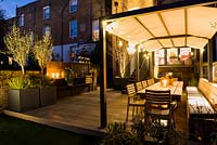 Illuminated outdoor kitchen and dining area with wooden tables and chairs and plants in containers including Olea europaea at night. 