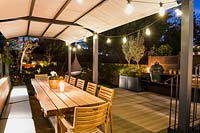Illuminated outdoor dining area with wooden tables and chairs and plants in containers including Olea europaea at night. 