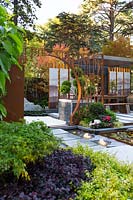 View across a garden planted in drifts of plants to a pavilion style Chinese garden, with a freestanding water feature, moongate, printed glass panels and a cloud pruned tree in a pot.