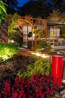 View across a water feature at night to moongate and a timber slab bench with steel legs, a bonsai conifer tree, showing spot lighting, underlighting featuring a red lantern.