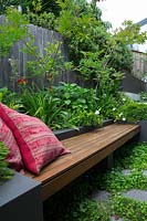 A cement rendered planter box with a hardwood timber inbuilt bench seat with two red patterned cushions, planted with Baby Panda Grass and a flowering Gardenia.