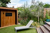London contemporary garden - border with Betula Utilis, olive tree, Stipa tenuissima next to wooden steps leading to upper patio area. A modern sun lounger is positioned on artificial turf lawn, with wooden gym or garden room