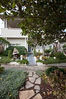A large sculptural sandstone boulder in the front garden of a house surrounded by Pride of Maderia, three totem pole art works, a clipped rosemary plant, the garden has potted succulents in the nature strip garden are bromelaids and succulents.