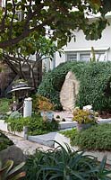 A large sculptural sandstone boulder in the front garden of a house surrounded by Pride of Maderia, the garden has potted succulents and bespoke art.