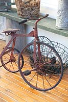 Detail of an old rusty children's tricycle on a timber verandah in front of a timber shelf with a collection of pots and wire baskets.