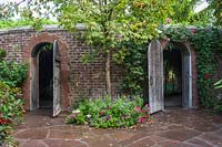 Brick walls with stone arches with wooden gates. Wall has Rosa - Climbing Rose over entrance and Aesculus glabra - Ohio Buckeye - underplanted between the gates